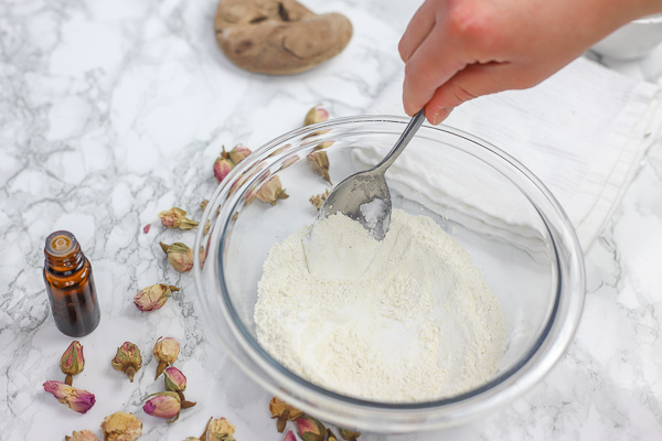 glass bowl with ingredients to make DIY deodorant being stirred with dried roses scattered around