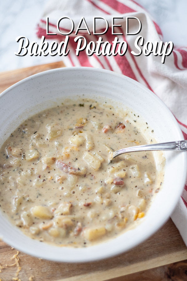 bowl of baked potato soup with bacon on a wood cutting board with a red and cream napkin to the right.