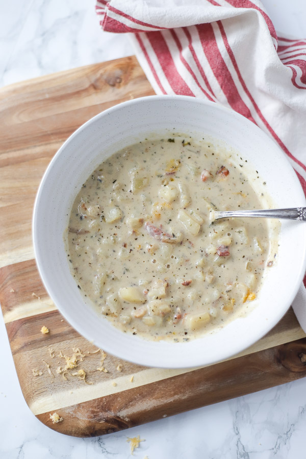 loaded baked potato soup in a cream bowl with a spoon. Bowl is on a wooden cutting board with a red and cream towel