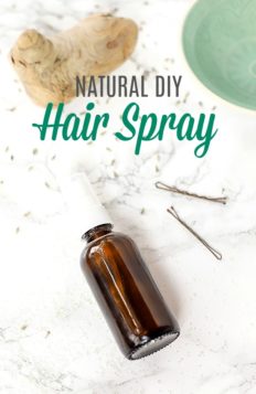 natural diy hair spray in amber bottle laying on a marble counter with bobby pins to the right, and a green bowl in the top right corner, and a piece of drift wood in the left corner