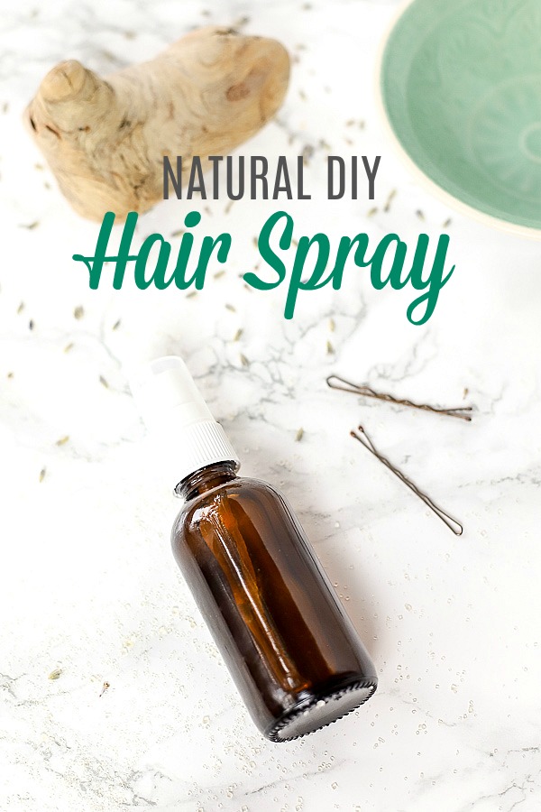 natural diy hair spray in amber bottle laying on a marble counter with bobby pins to the right, and a green bowl in the top right corner, and a piece of drift wood in the left corner