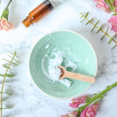 Baking soda face scrub in a small teal bowl with a wooden spoon. The bowl is surrounded by flowers and a bottle of essential oil.