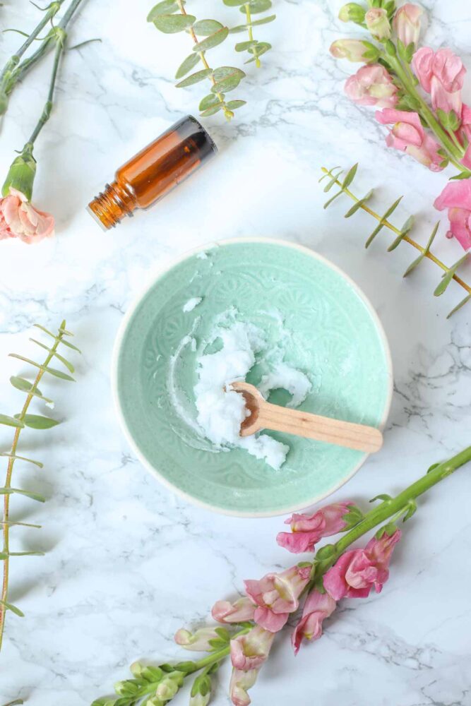 Baking soda face scrub in a small teal bowl with a wooden spoon. The bowl is surrounded by flowers and a bottle of essential oil.