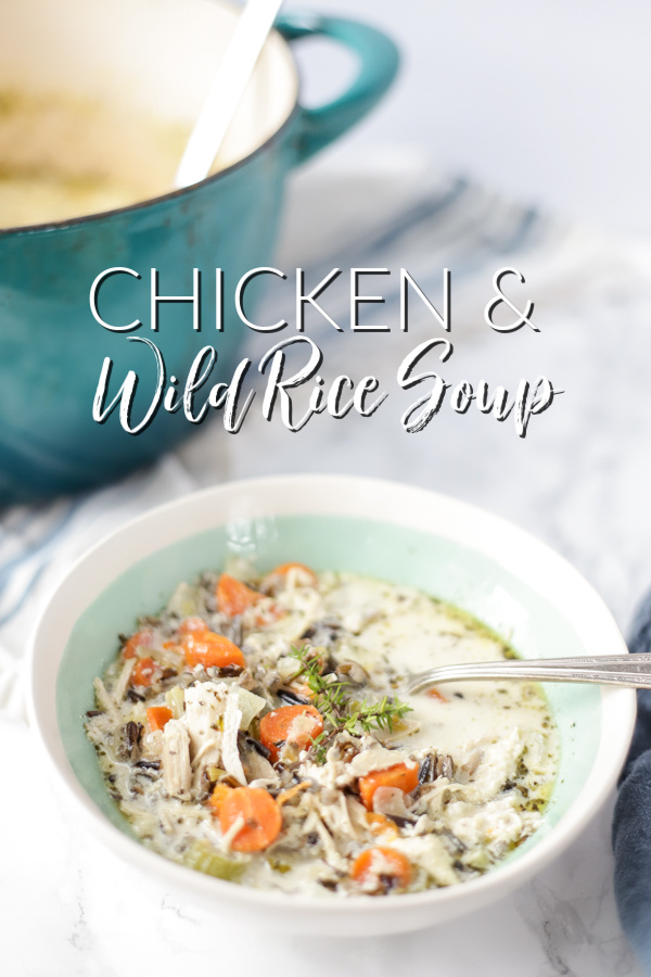creamy chicken and wild rice soup with carrots, celery, chicken, wild rice and onions in a creamy base and topped with fresh herbs in a cream and teal bowl with a dutch oven behind