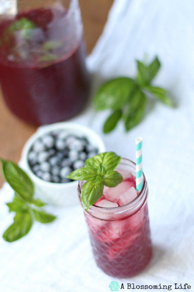 Blueberry Lemonade in a mason jar topped with basil. Frozen blueberry in a white dish behind it