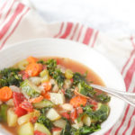 hearty bowl of minestrone soup with bacon, kale, potatoes, and other vegetables in chicken stock with an antique spoon in the bowl. Bowl is resting on top a red and tan napkin
