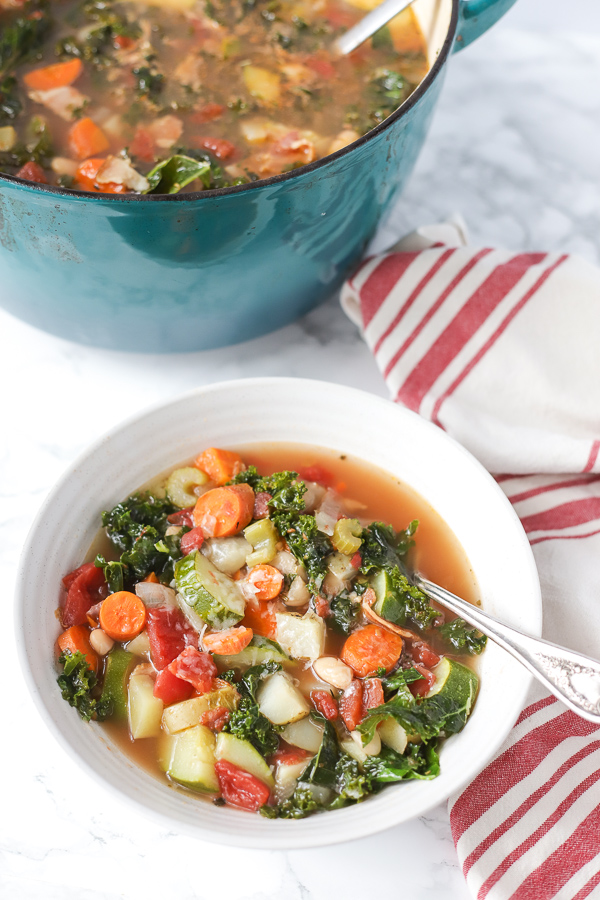 white bowl full of minestrone soup with an antique soon next to a red and tan napkin and a pot of minestrone soup behind it