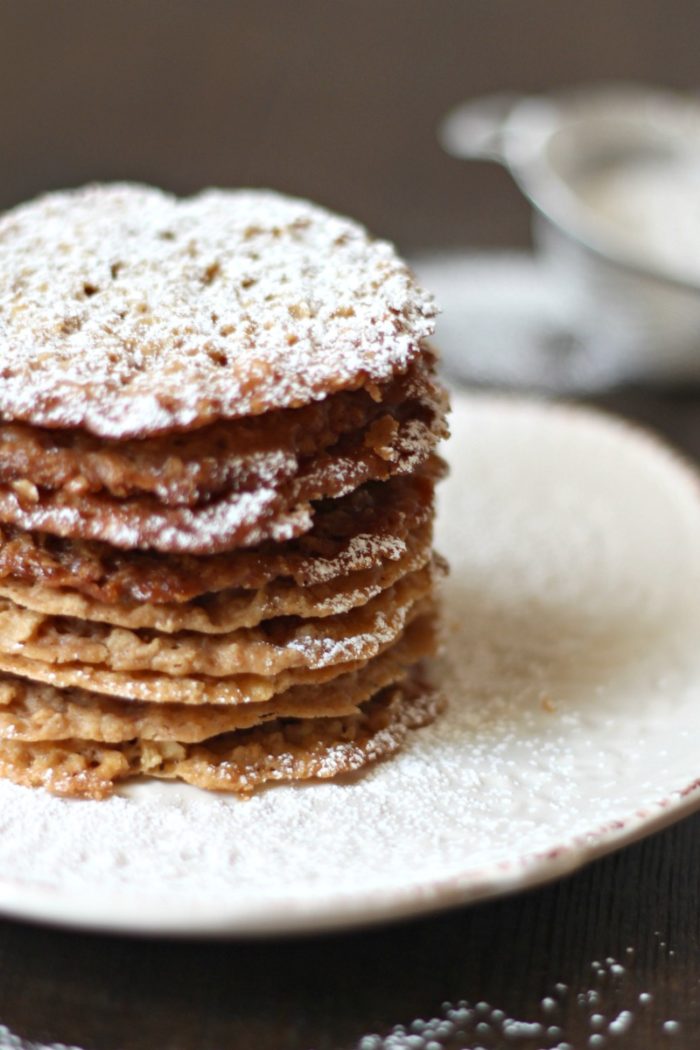 Oatmeal Lace Cookies stacked on a cream colored dish with sifter behind
