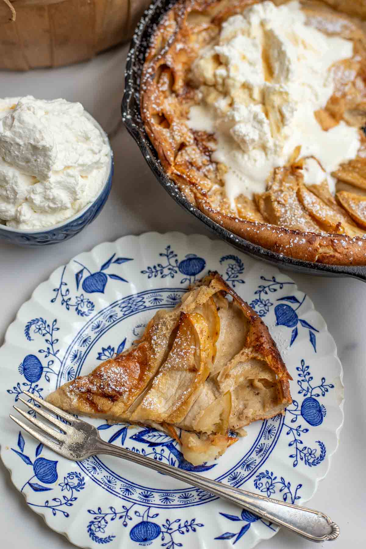 Slice of German apple pancake on a white and blue vintage plate with more of the dutch baby in the background and a bowl of whipped cream.
