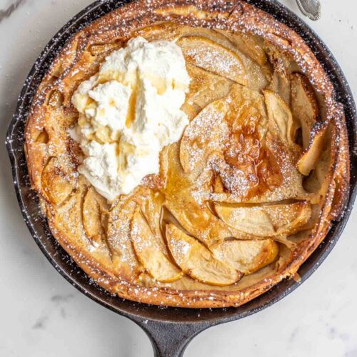 German apple pancake topped with powdered sugar, whipped cream, ad maple syrup in a cast iron skillet.