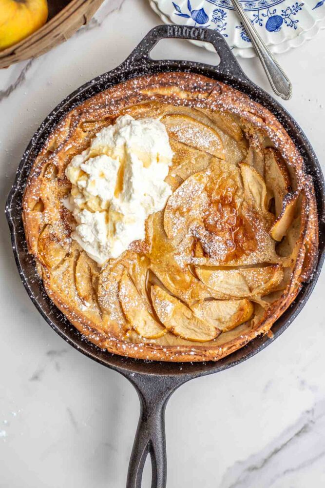 German apple pancake topped with powdered sugar, whipped cream, ad maple syrup in a cast iron skillet.