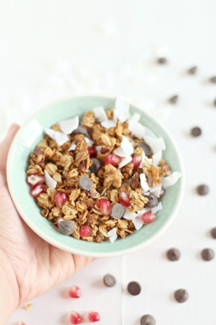 hand holding a teal bowl of homemade granola with chocolate chips, pomegranate seeds, and coconut shavings with chocolate chips and pomegranate seeds scattered around it 