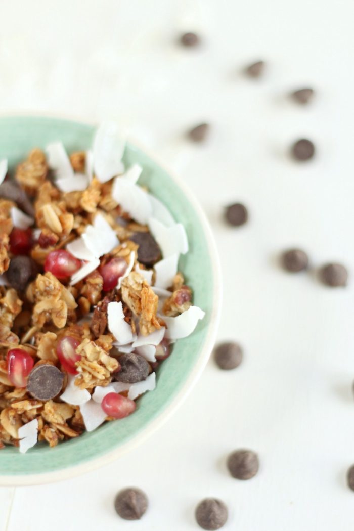 half teal bowl filled with homemade granola, chocolate chips, pomegranate seeds, and coconut shavings with chocolate chips scattered around the bowl