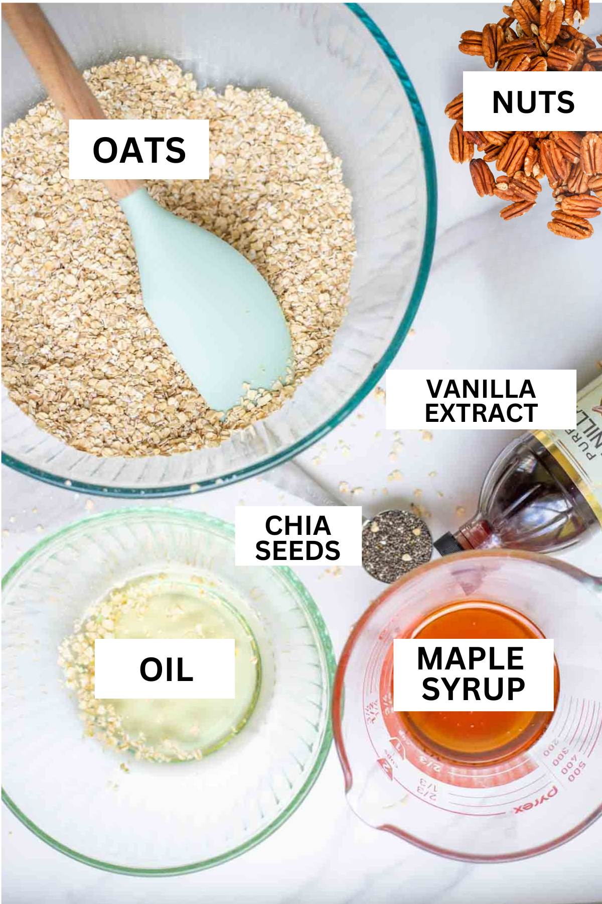 Bowls of oats, oil, maple syrup, with a bottle of vanilla extract, spoonful of chia seeds, and a pile of pecans on a countertop.
