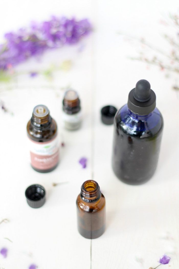 amber bottle of face serum with a blue bottle, essential oils, and purple flowers behind it.