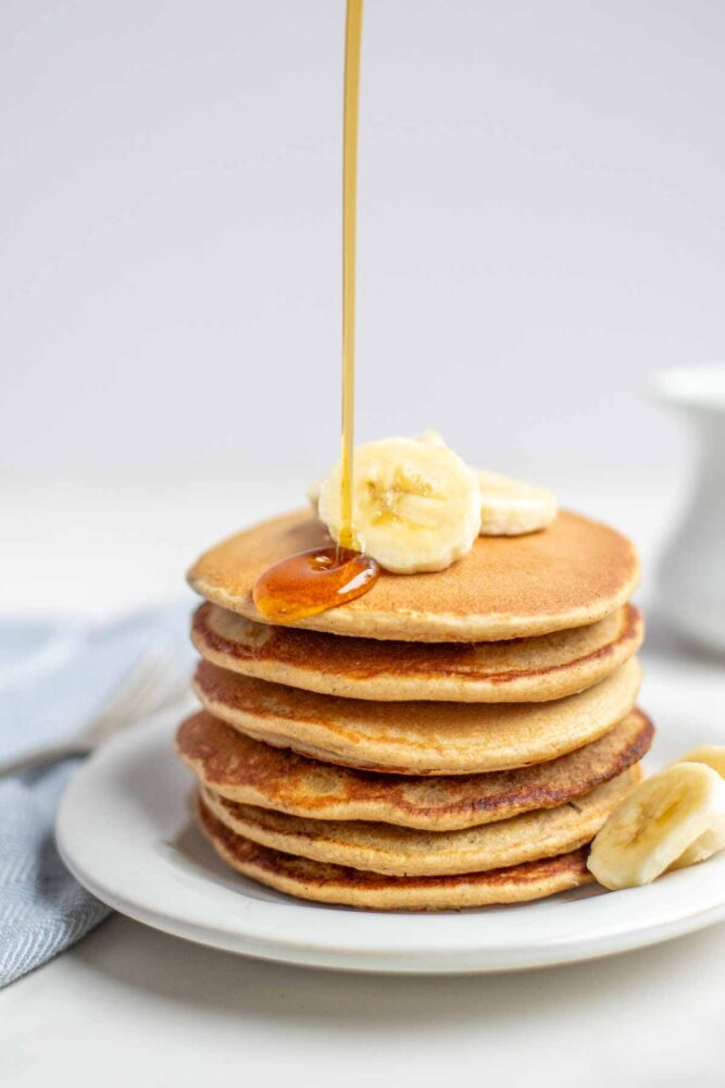 Banana oat pancakes in a stack topped with sliced bananas. Maple syrup is being poured on top of the pancakes.