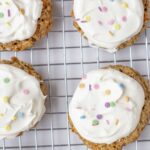 Carrot cake cookies topped with cream cheese frosting and sprinkles on a cooling rack.