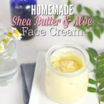 Shea butter cream sitting on a white place on a blue napkin with leaves and flowers in a jar behind it.