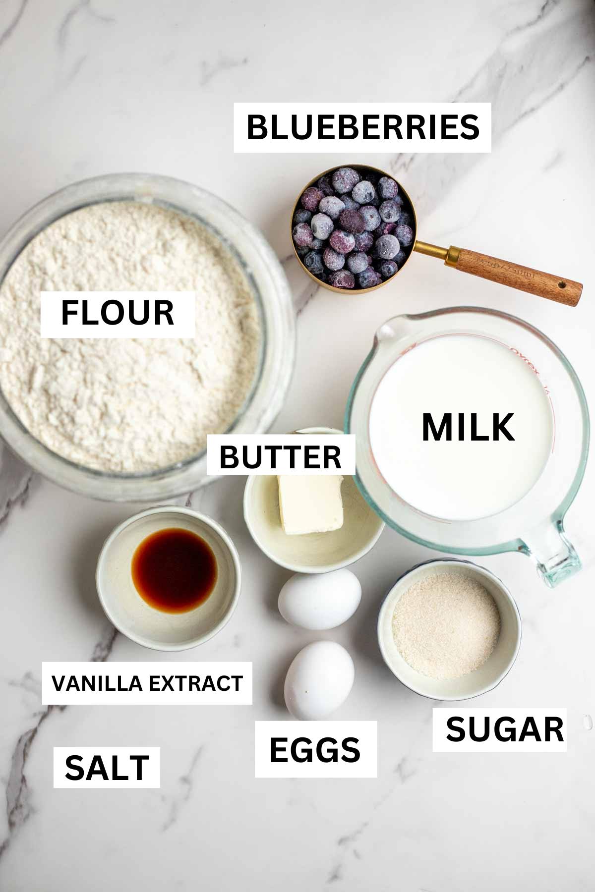 Jars and bowls of flour, milk, sugar, eggs, butter, vanilla extract, etc on a countertop.