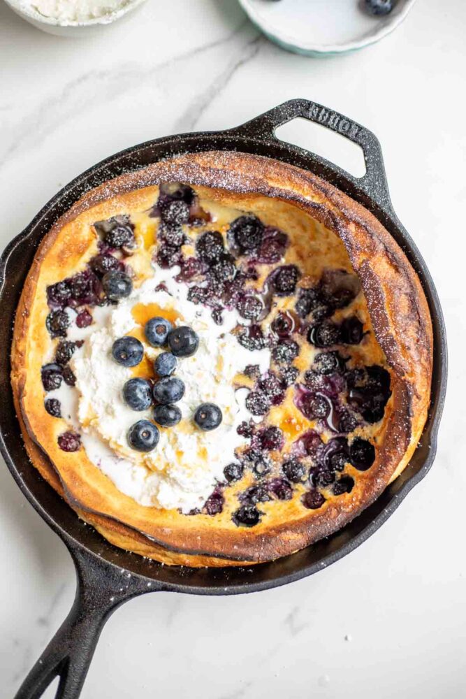 Blueberry dutch baby pancake topped with whipped cream, blueberries, maple syrup and powdered sugar in a cast iron skillet.