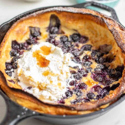 Blueberry dutch baby topped with whipped cream and maple syrup in a cast iron skillet. A bowl of whipped cream and a small container of blueberries and raspberries are behind the skillet.