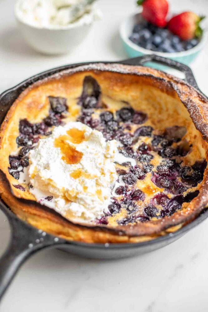 Blueberry dutch baby topped with whipped cream and maple syrup in a cast iron skillet. A bowl of whipped cream and a small container of blueberries and raspberries are behind the skillet.