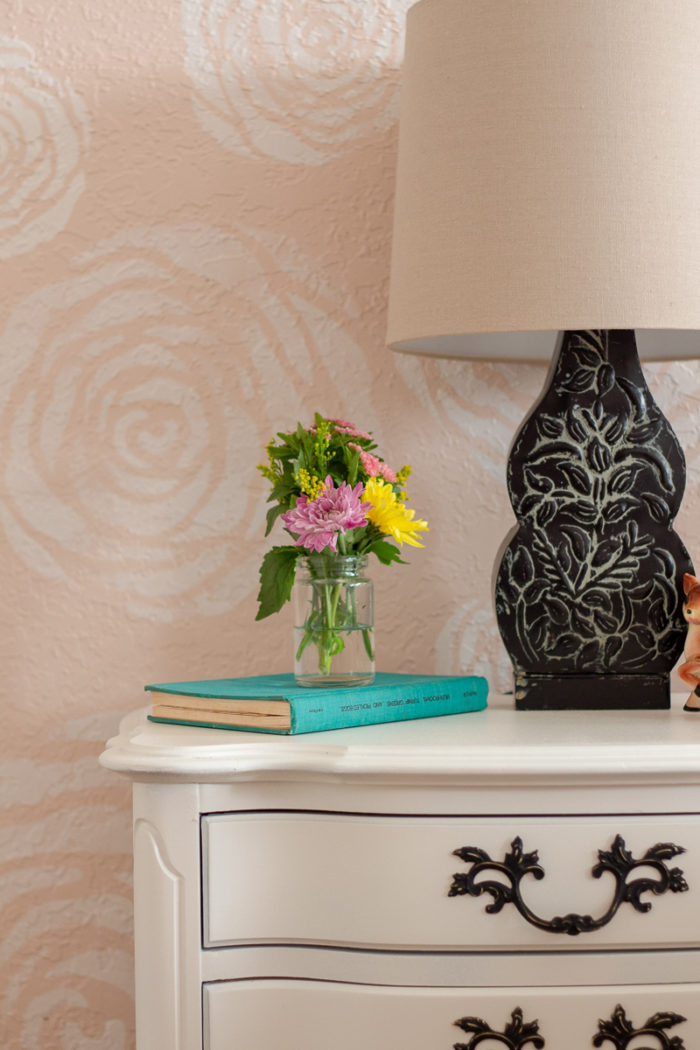 white painted antique nightstand topped with black lamp a turquoise book and mason jar filled with fresh flower in front of stenciled flower wall