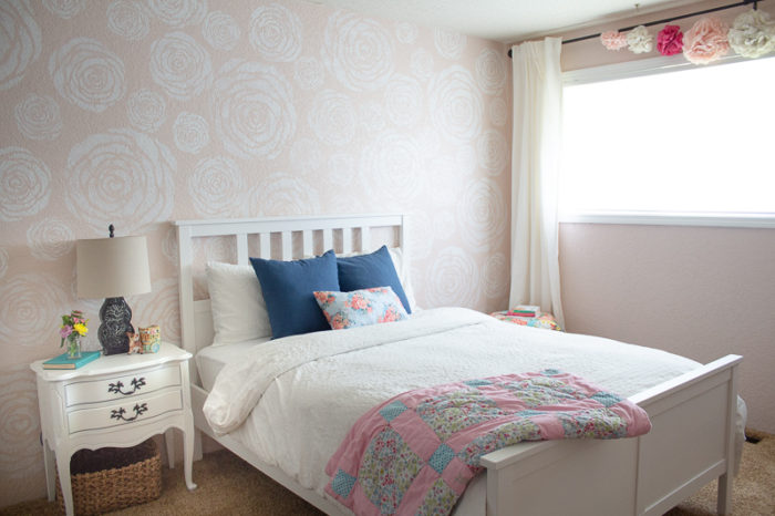 stenciled flowers on a wall with a white bed with white linens and blue pillow in front. An antique night stand to the left and a window with white linen curtains to the right