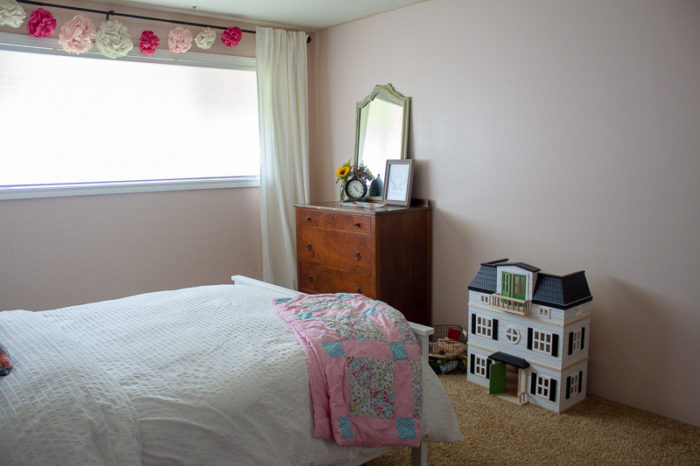 farmhouse style girls room with paper flower garland over window, antique dresser with antique green mirror. White magnolia dollhouse sits on the floor