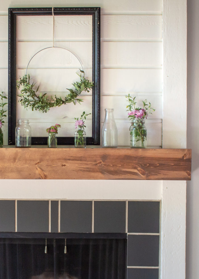 front half view of DIY fireplace mantel with stained wood and decorated with glass bottles, flowers, and a wreath- How to Build A Fireplace Mantel