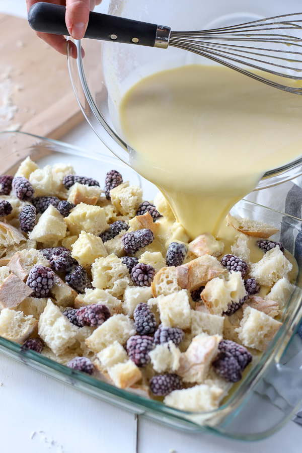 Baking dish with bread and blackberries with custard being poured over.