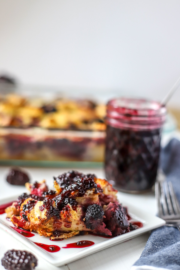 Slice of French Toast Casserole with Blackberries covered with blackberry syrup and a baking dish and jar of blackberry syrup in the back
