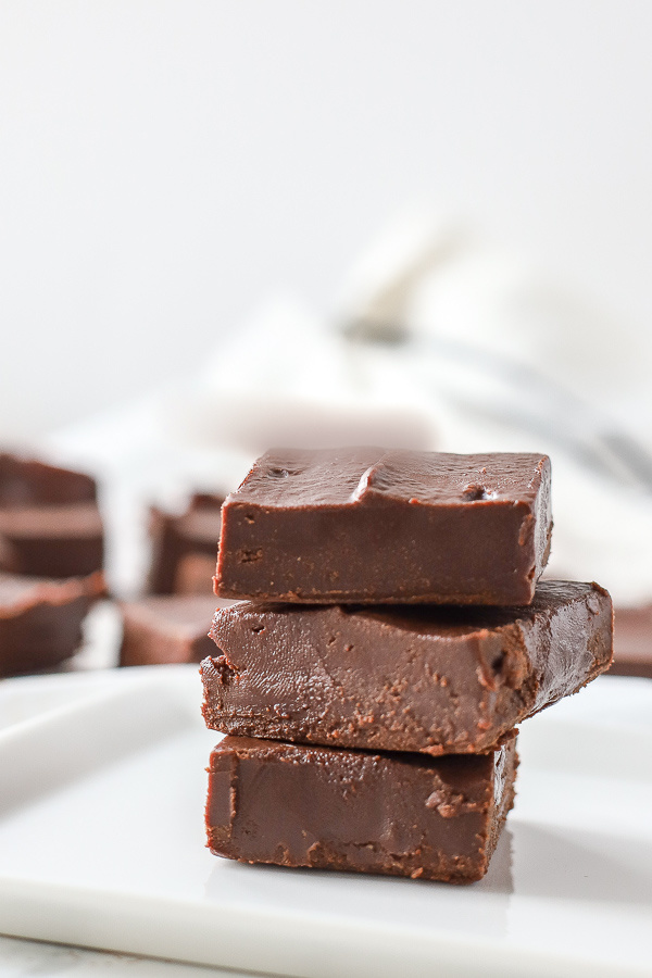 vegan fudge stacked 3 tall on a white plate with a white and black towel and more fudge pieces behind it