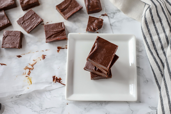 simple fudge recipe stacked on a white plate on a marble countertop with a white and black striped towel to the left and more fudge behind it