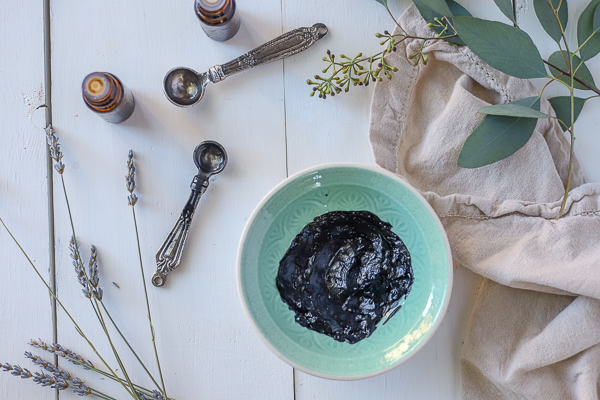 homemade activated charcoal mask in a teal bowl with measuring spoons and dried herbs around it.