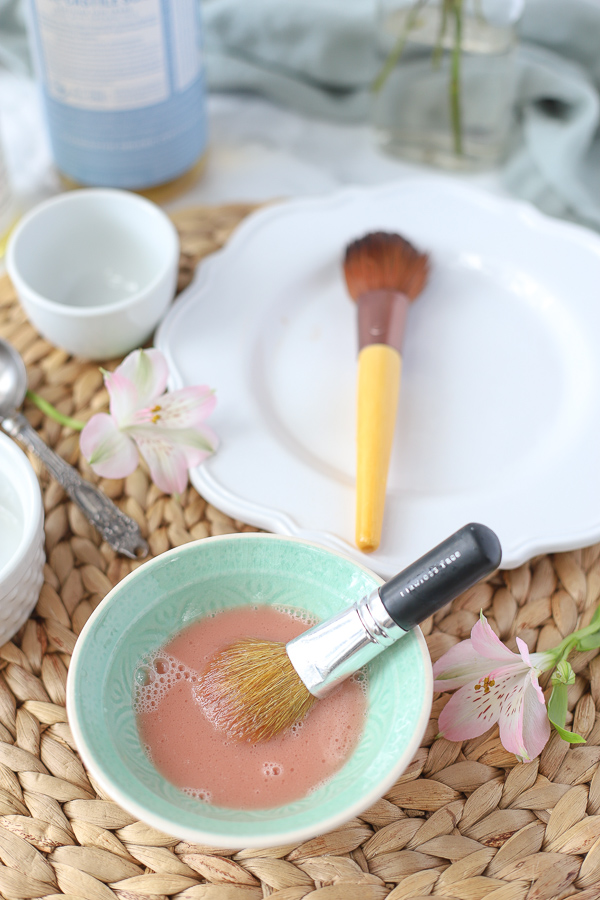 DIY makeup brush cleaner in a teal bowl with a dirty makeup brush in it and a white place with a clean makeup brush behind it