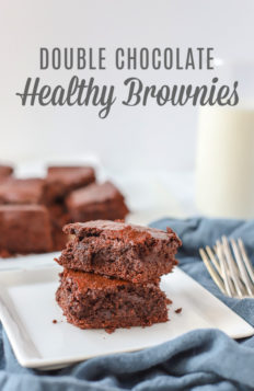 two double chocolate healthy brownies stacked on a white plate on top a blue napkin with a glass of milk and more brownies behind it
