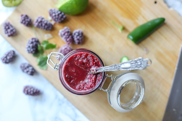 overhead photo of blackberry enchilada sauce in a glass container with a antique spoon coming out on top a wood cutting board with ingredients around it