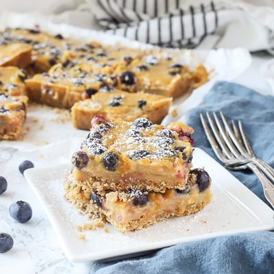 blueberry lemon bars stacked on a white plate with more dessert bars behind them