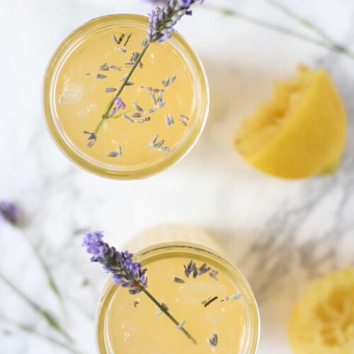 Overhead photo of two glasses of lavender lemonade with fresh sprigs of lavender int he glasses. The glasses are surrounded by a halved lemon and more lavender.