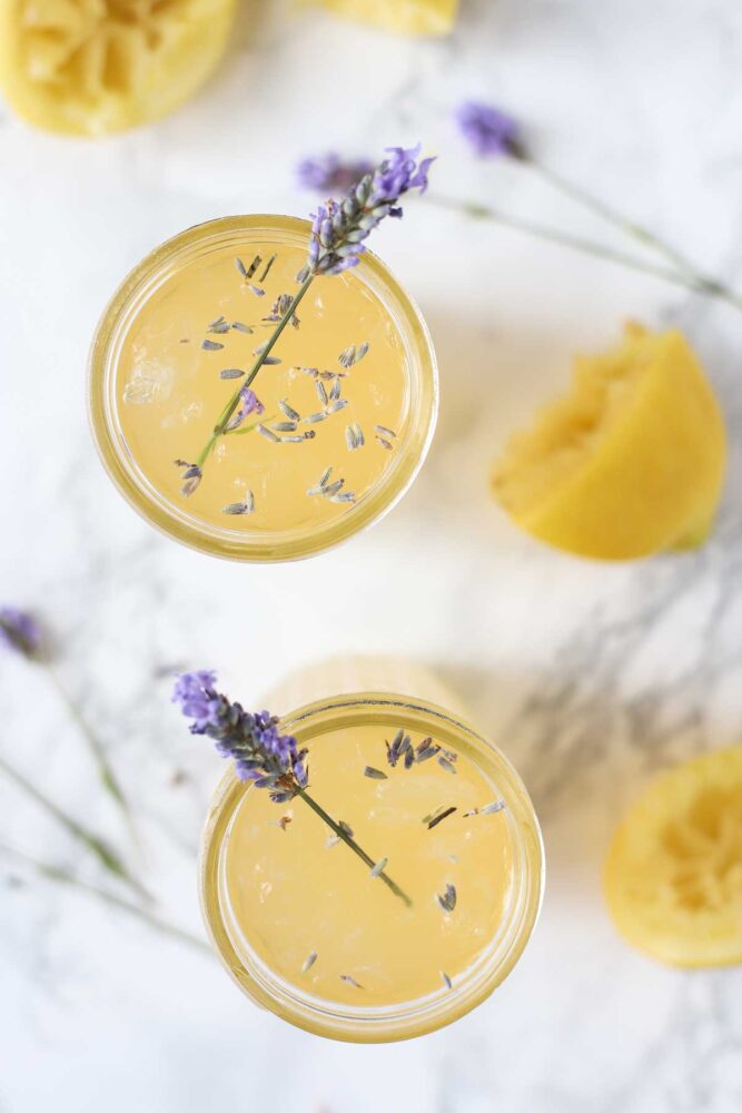 Overhead photo of two glasses of lavender lemonade with fresh sprigs of lavender int he glasses. The glasses are surrounded by a halved lemon and more lavender.