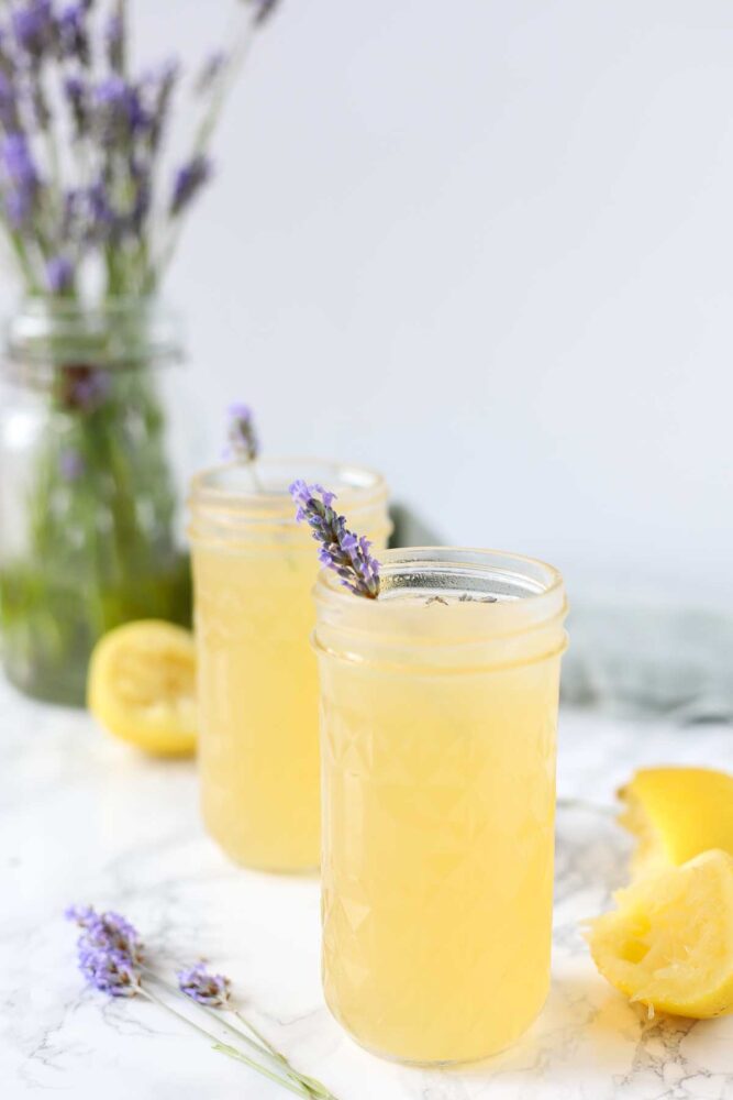 Two glasses of lavender lemonade with lavender sprigs in the glasses and a bunch of lavender in a vase in the background.