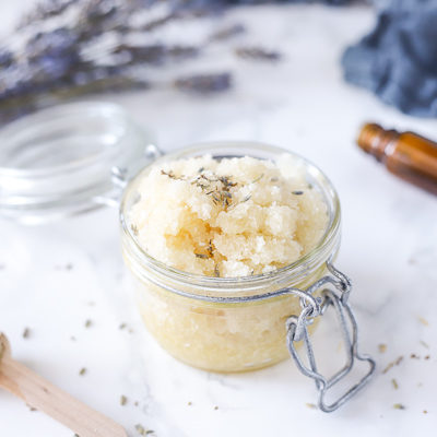 glass jar of exfoliating foot scrub with dried lavender buds sprinkled on the top. Bunch of lavender and blue napkin behind the jar