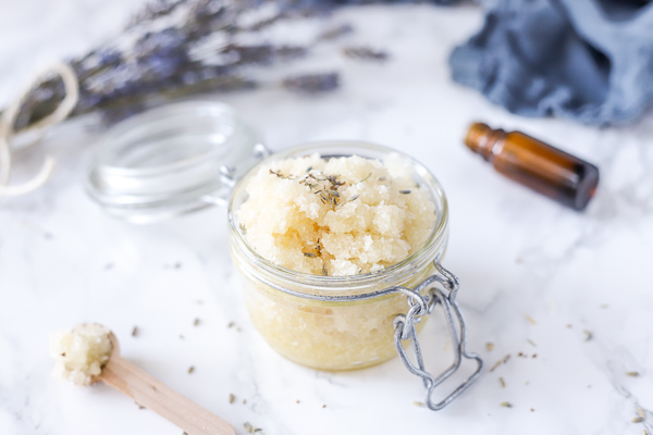 glass jar of exfoliating foot scrub with dried lavender buds sprinkled on the top. Bunch of lavender and blue napkin behind the jar