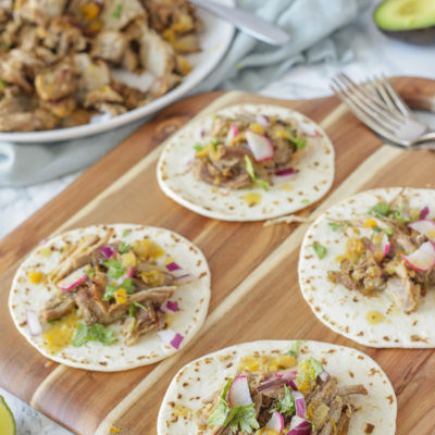 four pork carnitas topped with cilantro, radishes, and a squeeze of lime on a wood cutting board with plate of pork behind