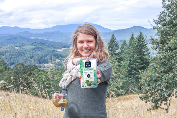 women in green sweater with the mountain in the background holding a box of teawell tea in one hand and a mug in tea in the other