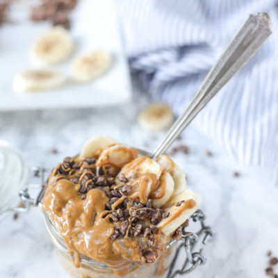 peanut butter overnight oats in a glass jar with a antique spoon sticking into the mixture. Bananas, peanut butter drizzle and chocolate chunks top the oatmeal