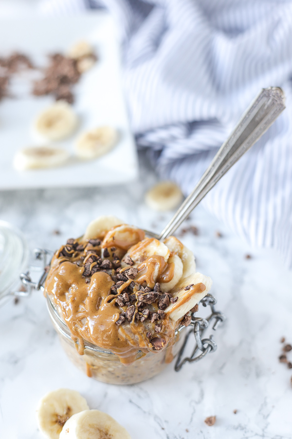 peanut butter overnight oats in a glass jar with a antique spoon sticking into the mixture. Bananas, peanut butter drizzle and chocolate chunks top the oatmeal