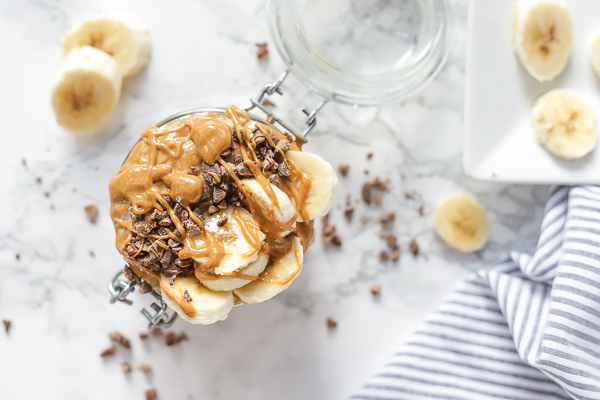 overhead shot of peanut butter overnight oats, topped with sliced bananas, chocolate crumbles, and drizzled with peanut butter. sliced bananas and a napkin are in the background