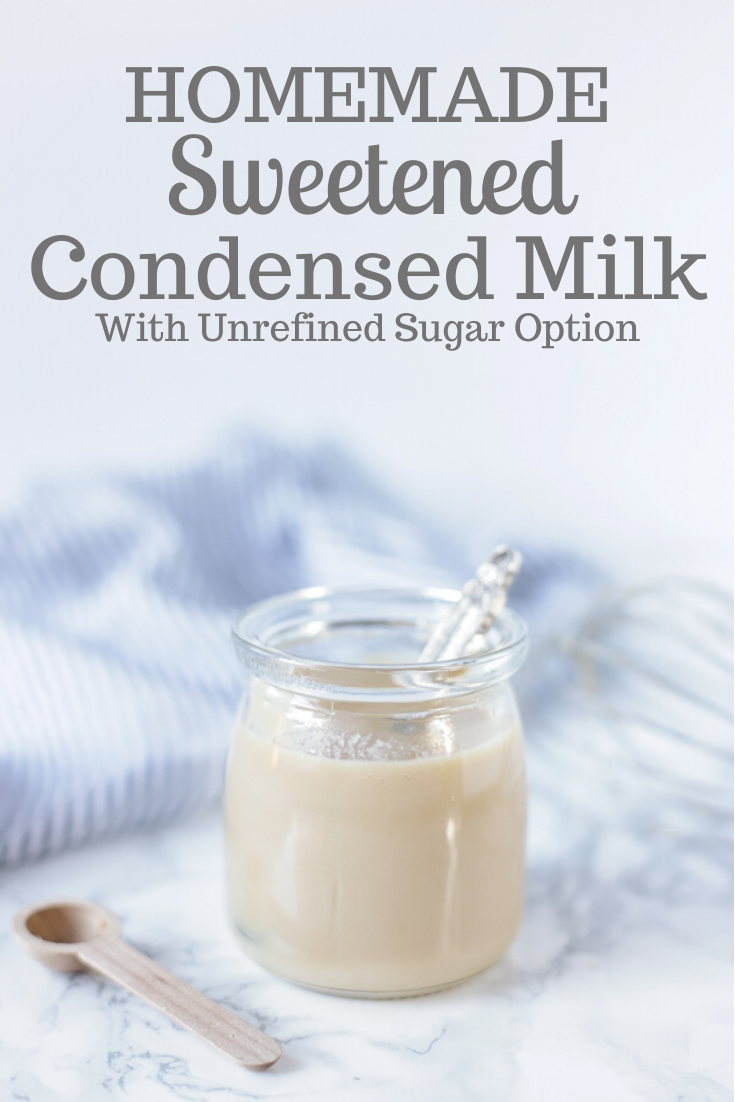 Homemade sweetened condensed milk in a glass jar with an antique spoon in it. A wooden spoon is sitting next to the jar and a blue and white stripped towel in the back